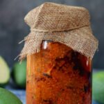 Stock photo showing close-up view of green (raw) mango chutney recipe in a glass jar covered with hessian on blue wood grain surface. Ingredients include, powdered turmeric, nigella onion seeds, red chilli powder, fenugreek seeds, pickling salt and mustard seeds.